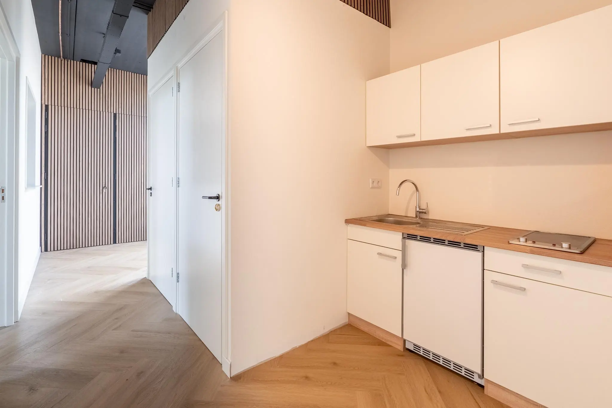 Kleine moderne kitchenette met witte kastjes en houten werkblad in een strak afgewerkte ruimte.