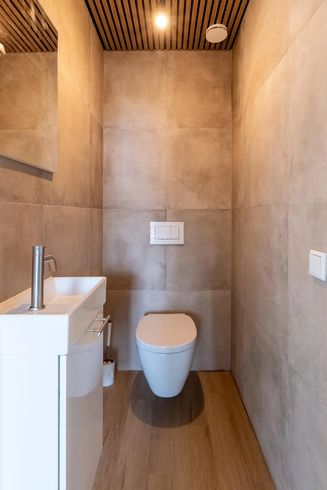 Modern, minimalistisch toilet met wandcloset, kleine wastafel en houten plafondaccenten.