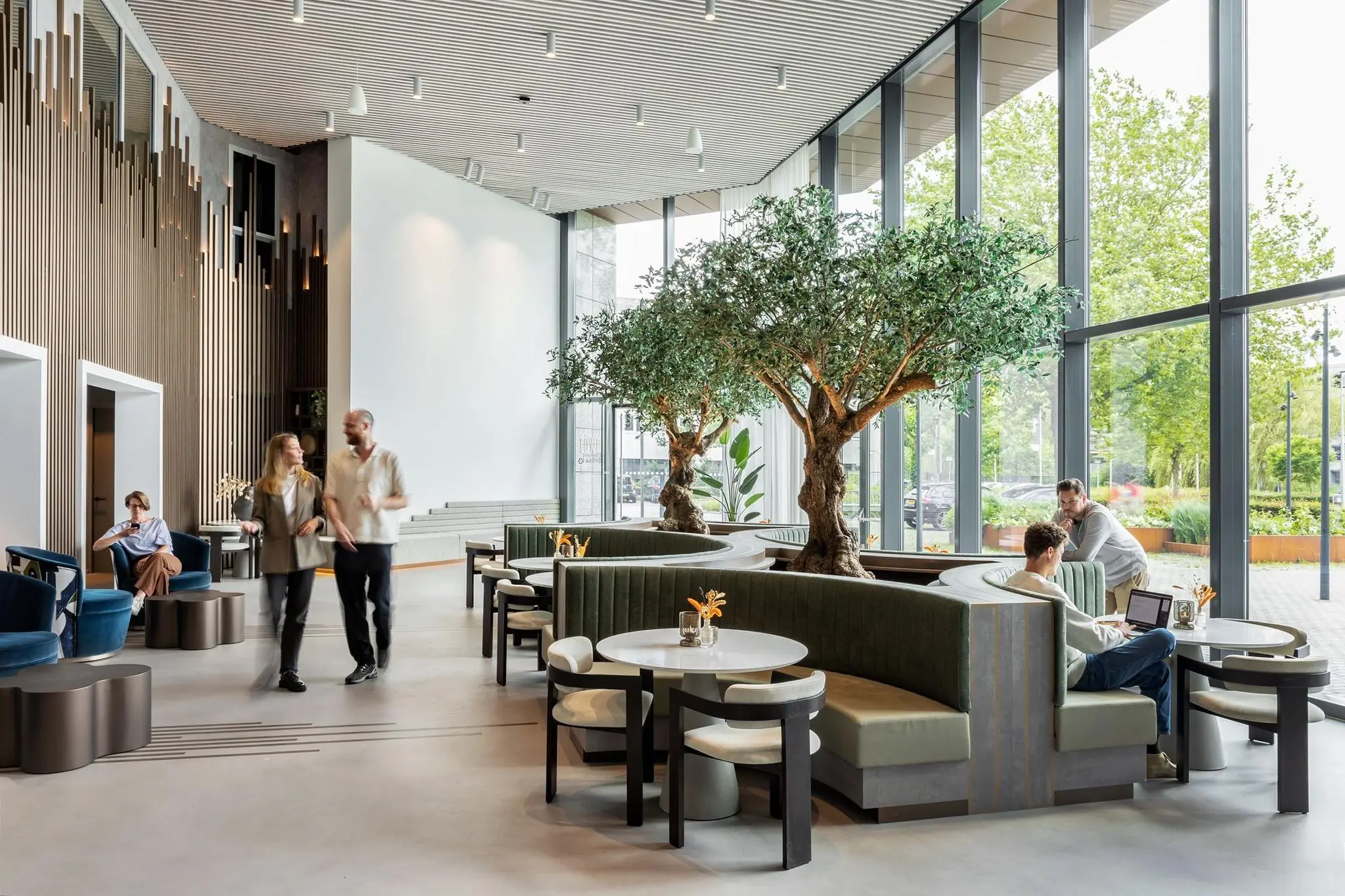 Moderne, open lobbyruimte in het Diemerhof met zitgedeelte, grote ramen en stijlvolle inrichting met olijfbomen.