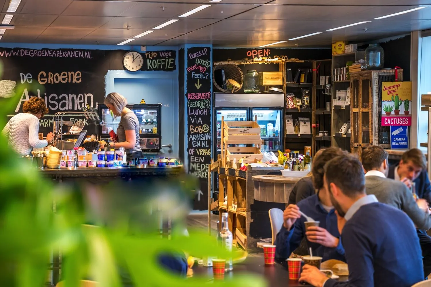 Mensen eten en drinken in lunchcafé De Groene Artisanen aan de Reactorweg, met zelfbedieningsbalie en kleurrijke inrichting.