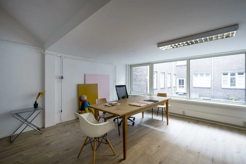 Licht en minimalistisch ingericht kantoor met houten vloer, grote ramen en een vergadertafel met diverse stoelen.