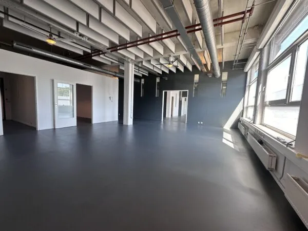 Lichte, industriële kantoorruimte aan de Pilotenstraat met grote ramen, donkere vloer en zichtbare plafondleidingen.