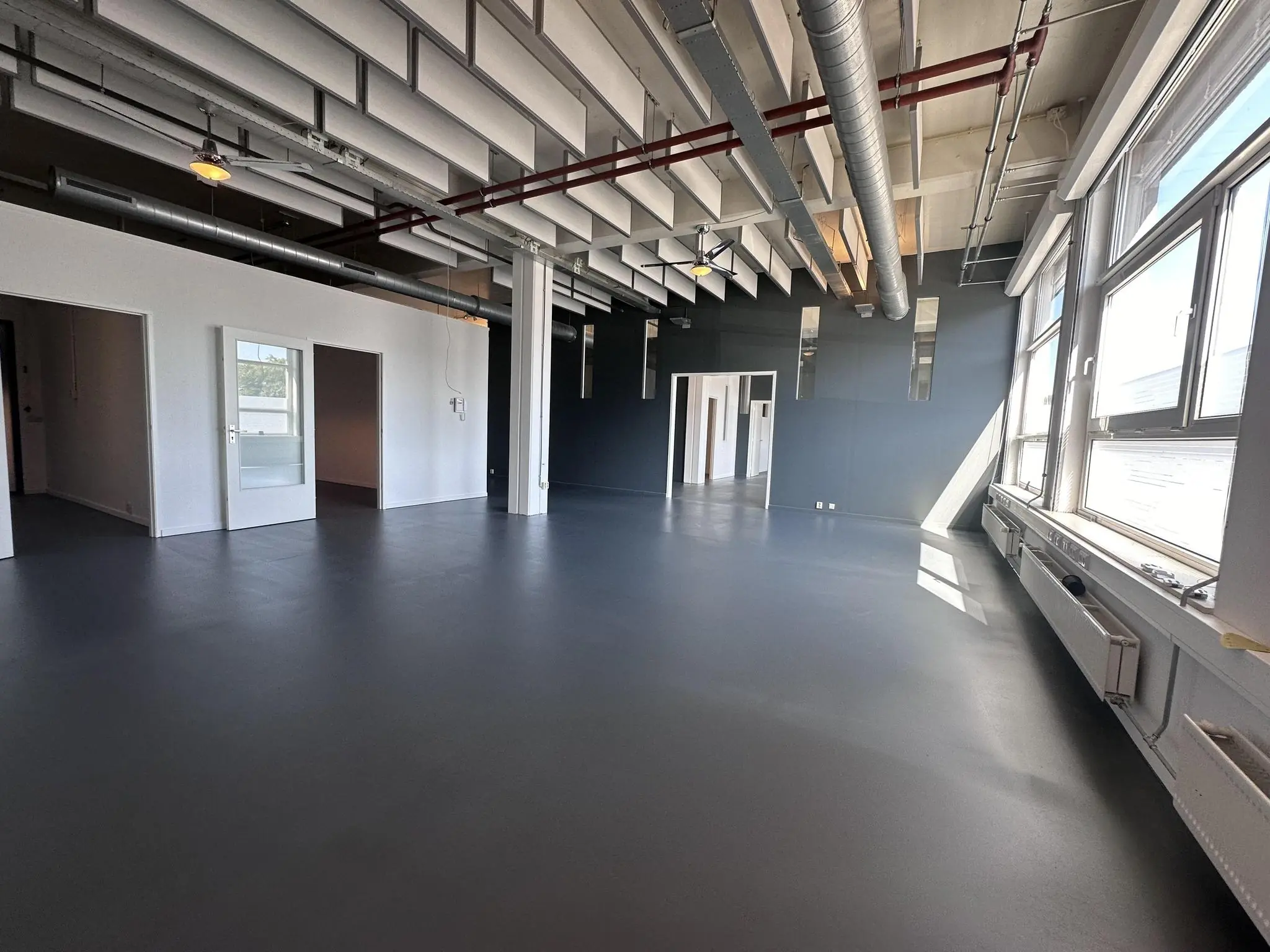 Lichte, industriële kantoorruimte aan de Pilotenstraat met grote ramen, donkere vloer en zichtbare plafondleidingen.