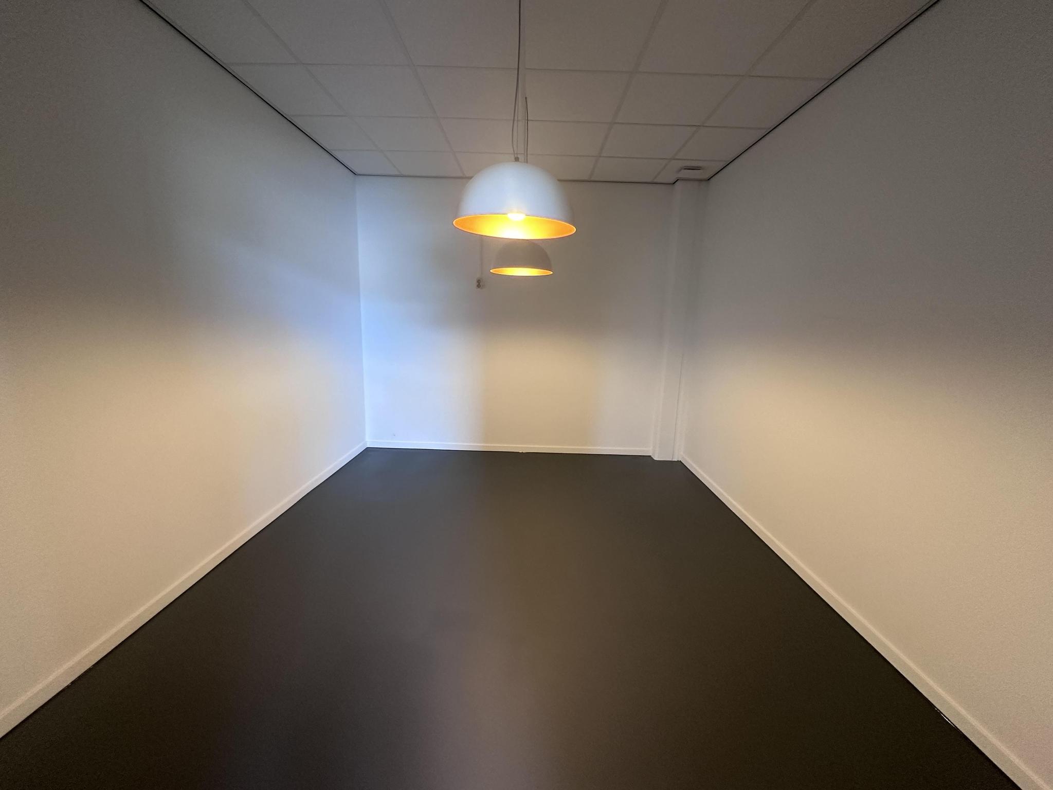 Lege kantoorruimte aan de Pilotenstraat met witte muren, donkere vloer en twee hanglampen aan het plafond.
