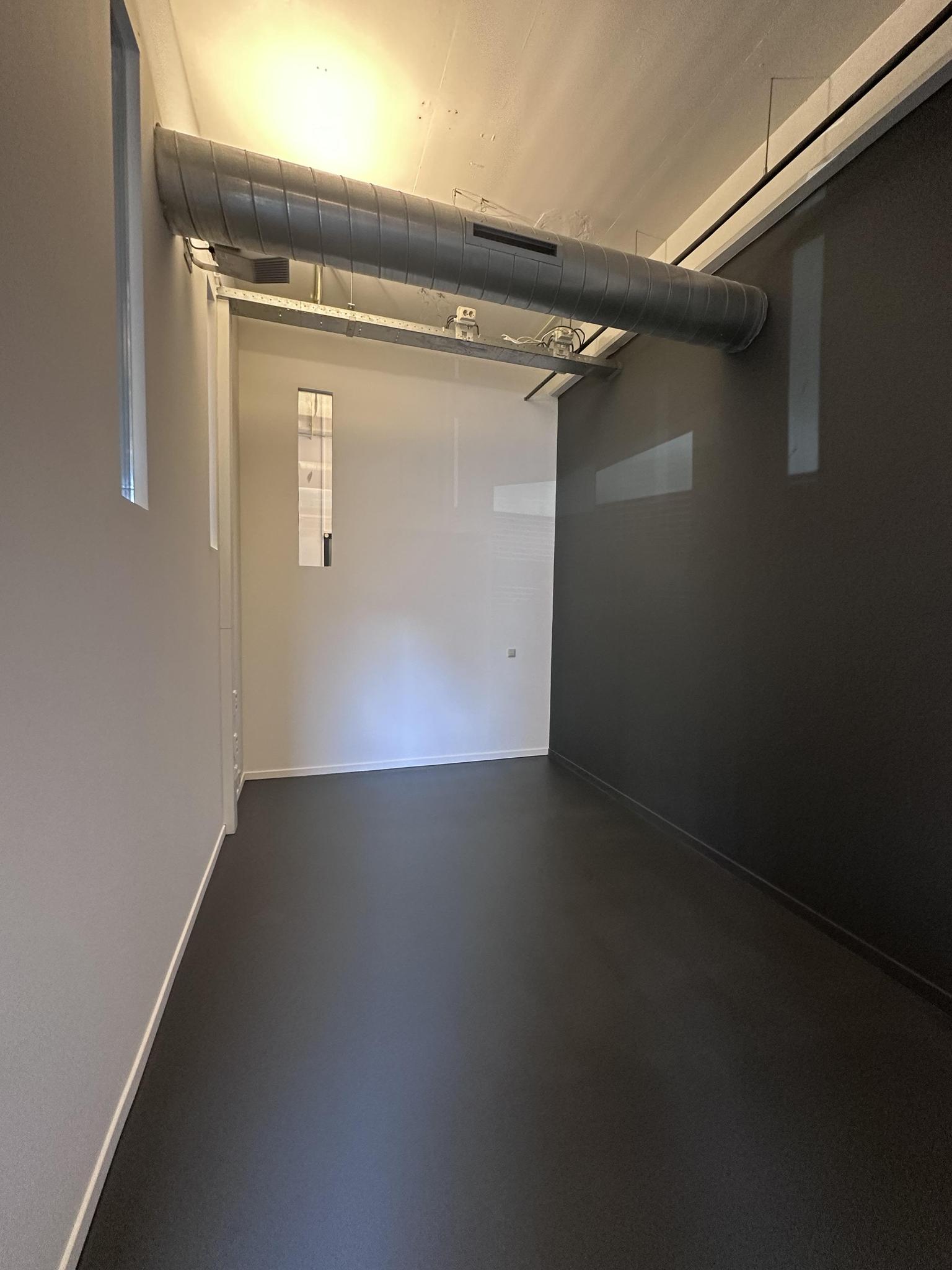 Lege, smalle ruimte aan de Pilotenstraat met een donkere vloer, witte en zwarte muren en zichtbaar ventilatiekanaal aan het plafond.