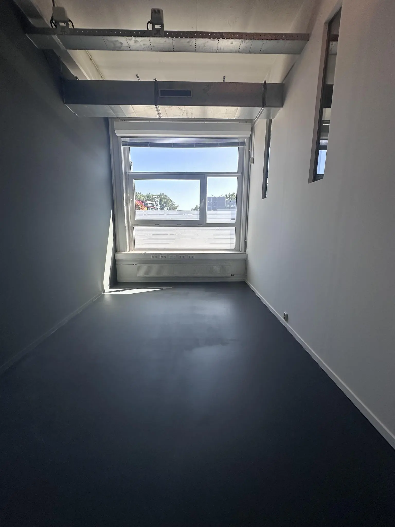 Lege kantoorruimte aan de Pilotenstraat met groot raam, donkergrijze vloer en zichtbaar ventilatiekanaal aan het plafond.