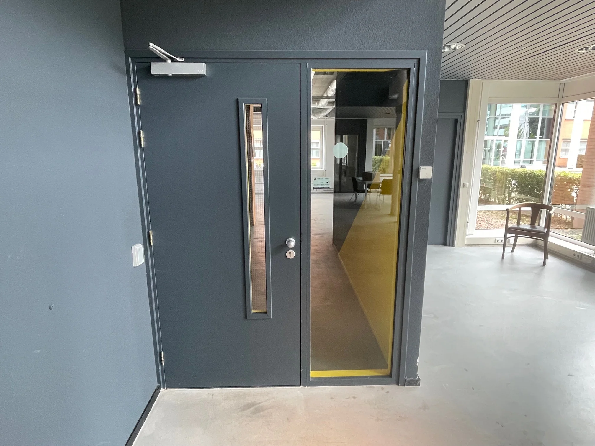 Zwarte deur met smal raam en daarnaast een glazen deur met gele rand, leidend naar een moderne gang in een kantoorgebouw.