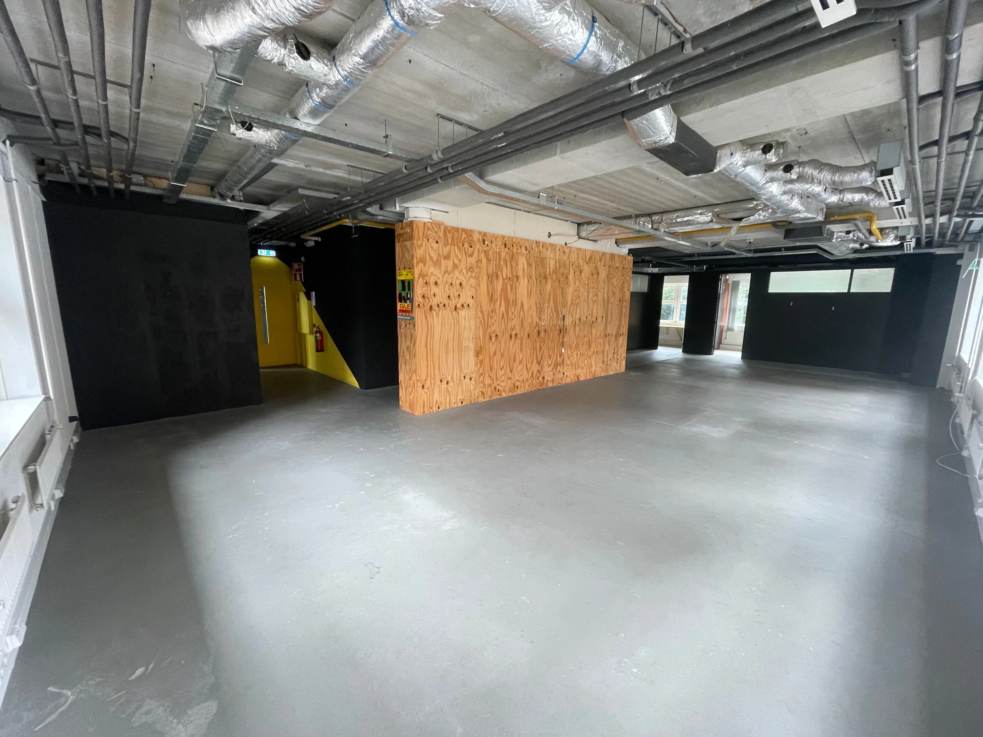 Industriële, lege ruimte met betonnen vloer, zichtbare leidingen aan het plafond en een houten scheidingswand.