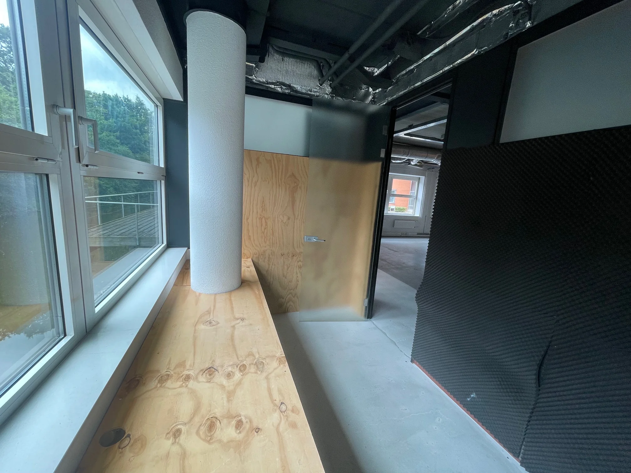 Smalle gang met houten afwerking, akoestisch schuim op de muur en zicht op een aangrenzende ruimte met ramen.