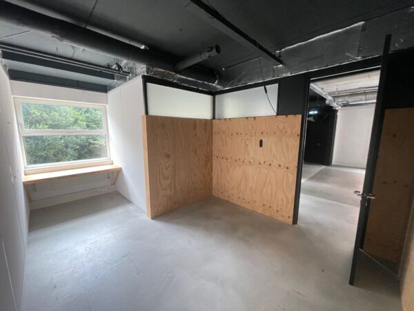 Kleine, industriële kantoorruimte met betonnen vloer, houten tussenschot en bureau bij het raam.