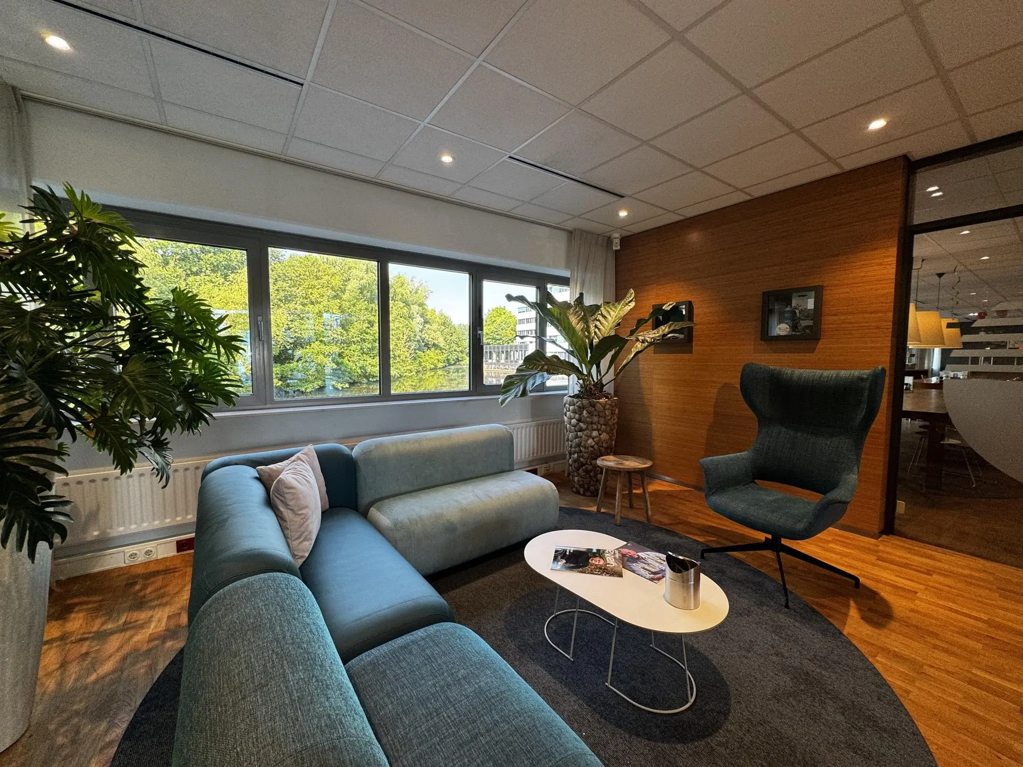 Moderne wachtruimte met een blauwgroene zithoek, planten en een groot raam met uitzicht op bomen.