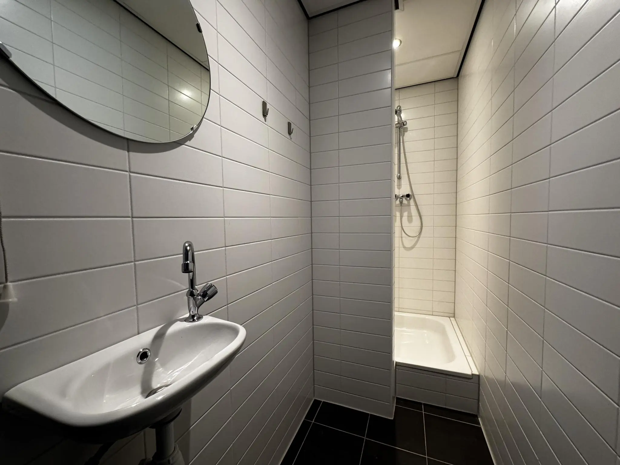 Kleine, wit betegelde badkamer met wastafel, ronde spiegel en inloopdouche.