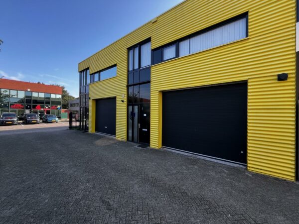Bedrijfsgebouw met gele gevel en zwarte garagedeuren aan de Ambachtsweg, met parkeerplaats en ander bedrijfspand op de achtergrond.