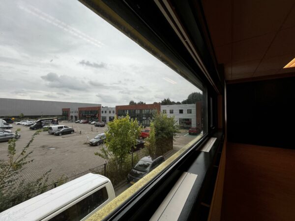 Uitzicht vanuit een raam op een bedrijventerrein aan de Ambachtsweg met geparkeerde auto's en bedrijfsgebouwen.