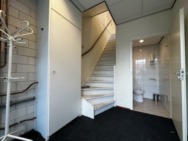 Ingangsruimte aan de Ambachtsweg met een trap, een openstaande deur naar een toilet en zichtbare leidingen langs de muur.