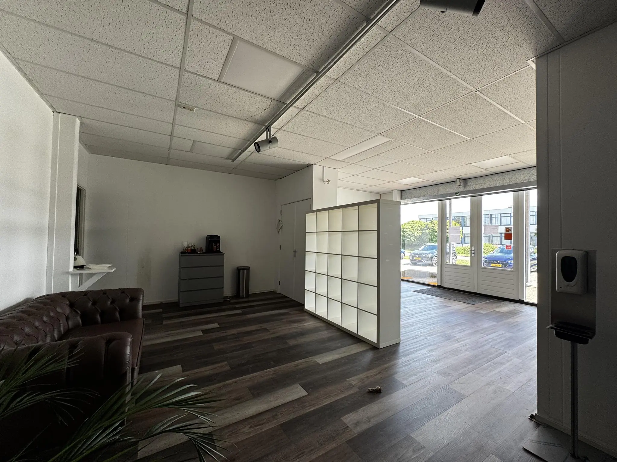 Ontvangsthal aan de Lorentzweg met open ruimte, een bruine leren bank, witte vakkenkast en zicht op de parkeerplaats buiten.