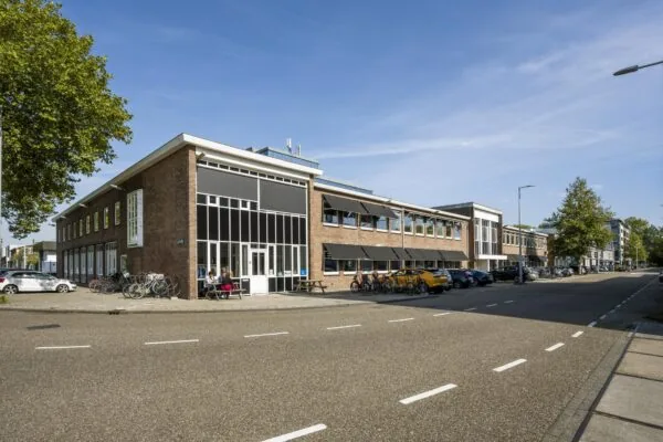 Bedrijfsverzamelgebouw aan de Vliegtuigstraat in een zonnige straat met geparkeerde auto's en fietsen.