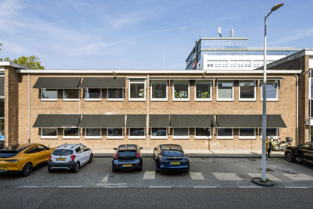 Voorgevel van een bakstenen kantoorgebouw aan de Vliegtuigstraat met geparkeerde auto’s en zwarte zonneschermen voor de ramen.