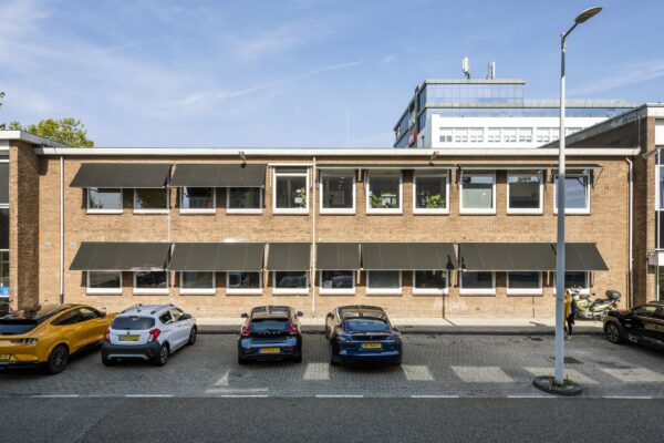 Voorgevel van een bakstenen kantoorgebouw aan de Vliegtuigstraat met geparkeerde auto’s en zwarte zonneschermen voor de ramen.