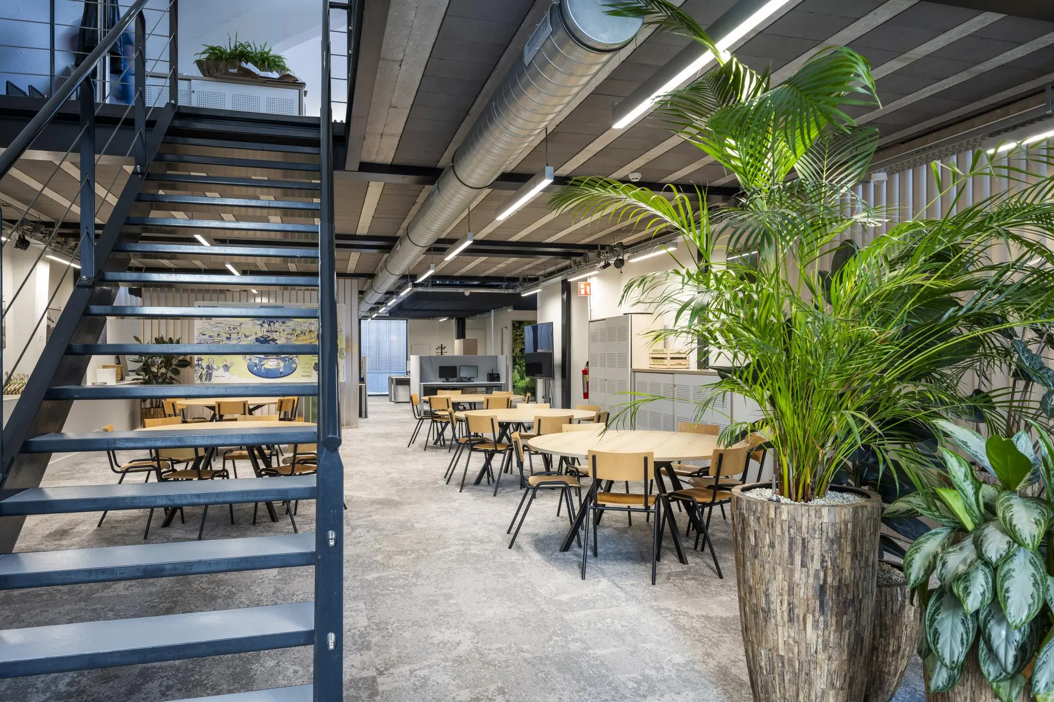 Moderne, open gemeenschappelijke ruimte met houten tafels, groene planten en een industriële trap aan de Abram Dudok van Heelstraat.