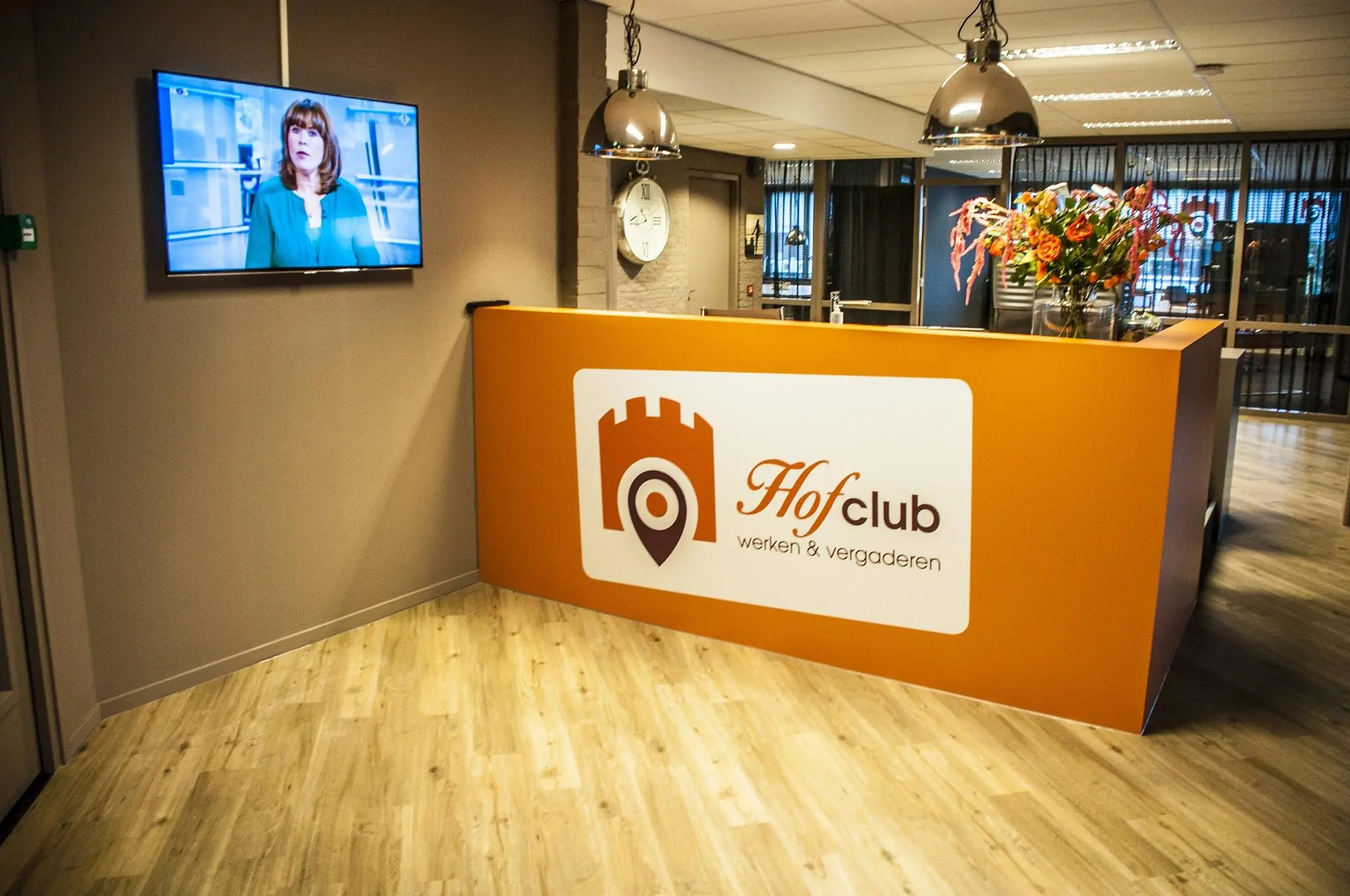 Moderne ontvangstruimte van Hofclub werken & vergaderen met een oranje balie, bloemenvaas en een televisiescherm aan de muur.