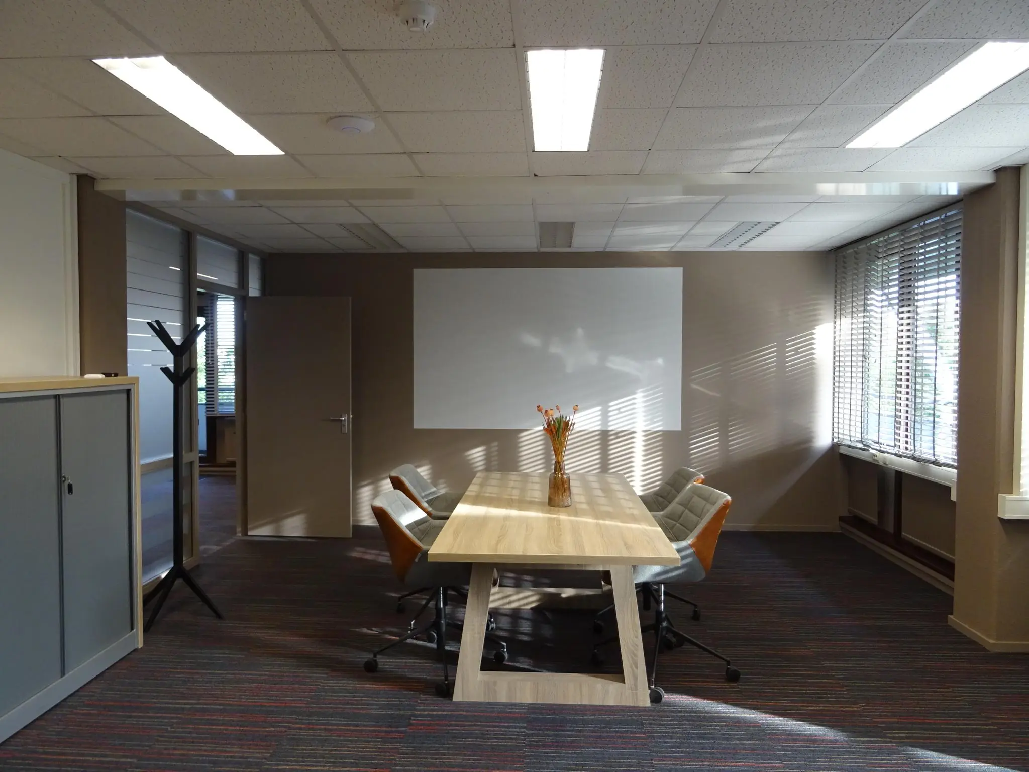 Vergaderruimte aan de Zaagmolenlaan met een houten tafel, zes stoelen, een whiteboard en zonlicht dat door jaloezieën naar binnen valt.