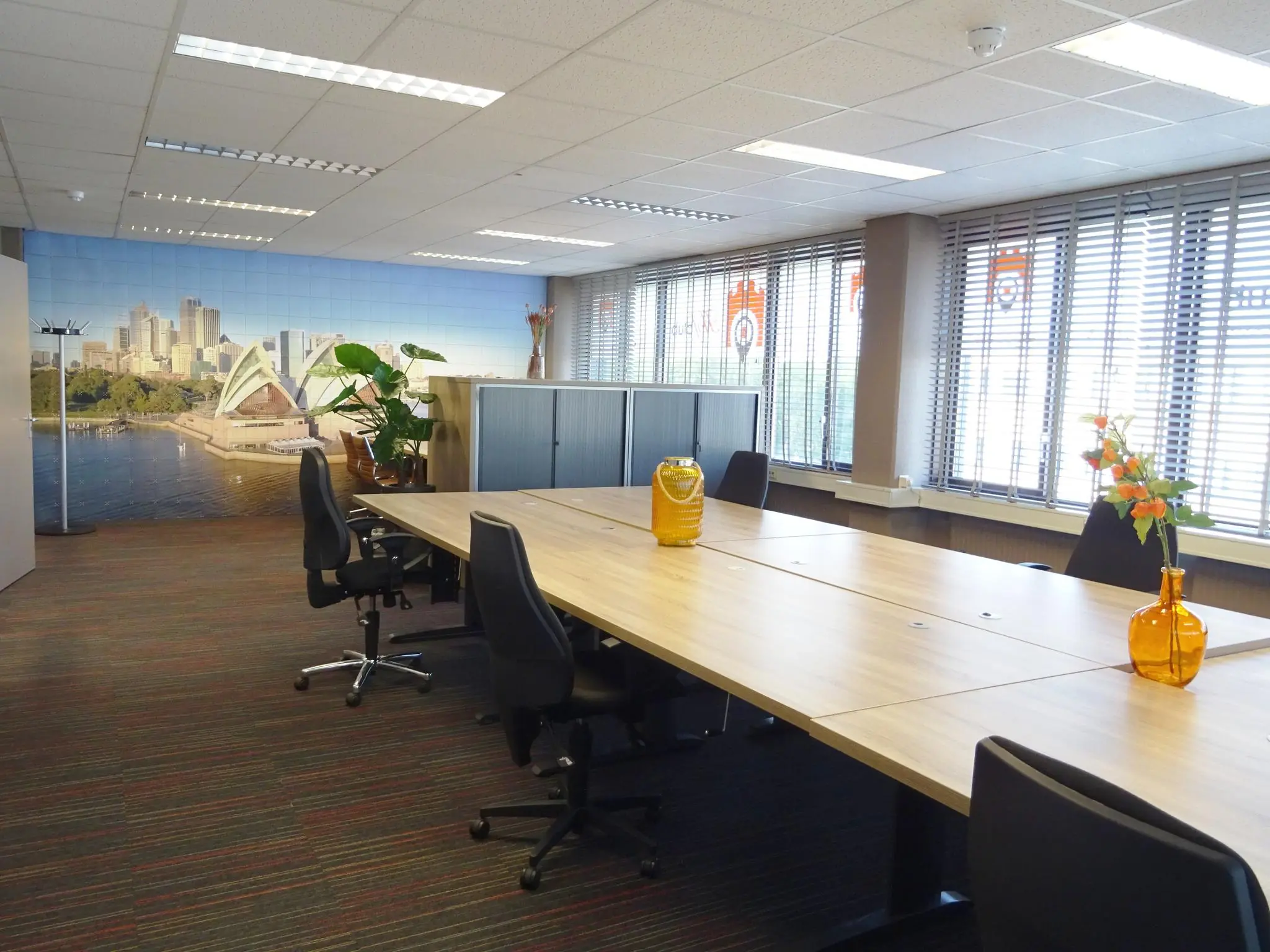 Modern kantoor met gedeelde werkplekken, oranje vazen en een muurfoto van de skyline van Sydney.