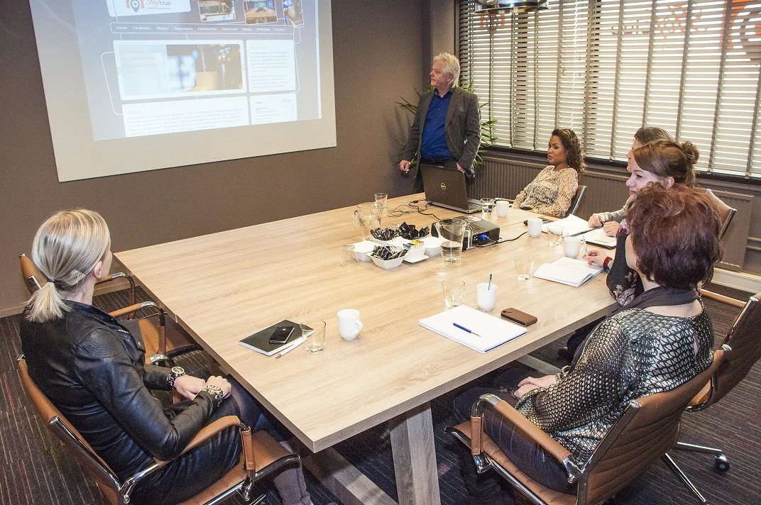 Vijf vrouwen en een man houden een presentatie in een vergaderruimte aan de Zaagmolenlaan, met een projectie op het scherm.