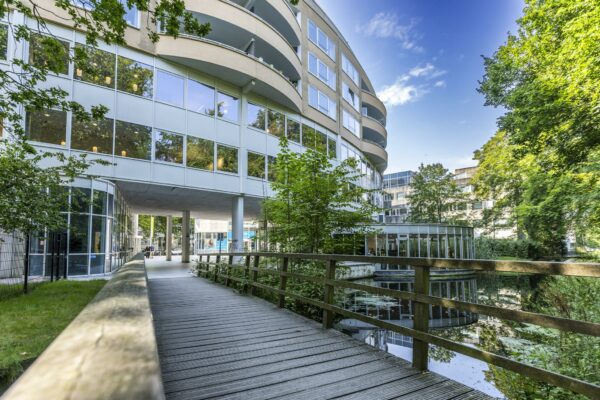 Modern kantoorgebouw aan de Bezuidenhoutseweg met een houten voetgangersbrug over het water en omliggende groene bomen.