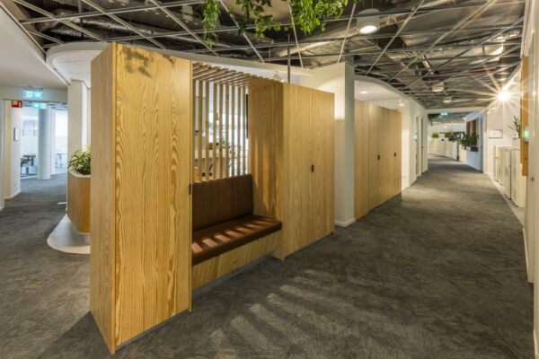 Moderne kantoorruimte aan de Bezuidenhoutseweg met houten zitnissen, grijze vloerbedekking en een open plafond met zichtbare leidingen en planten.
