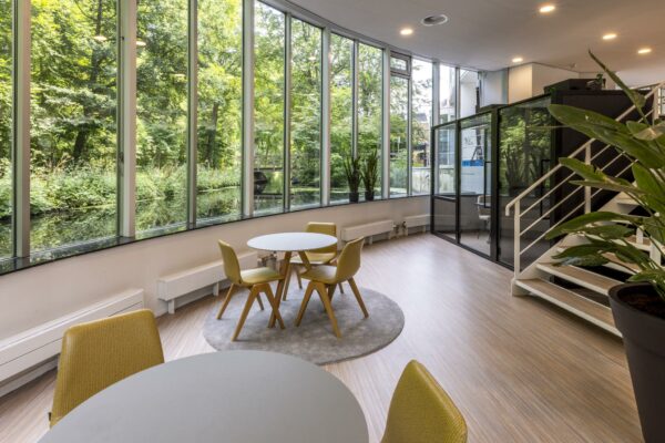 Moderne kantoorruimte aan de Bezuidenhoutseweg met grote ramen, uitzicht op groen en een zithoek met gele stoelen.