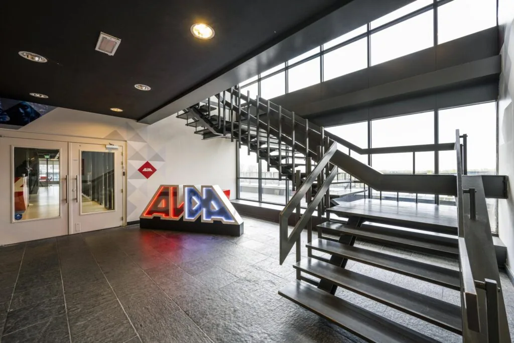 Moderne entreehal met stalen trap, grote raampartijen en een verlicht ALDA-logo bij Anthony Fokkerweg.