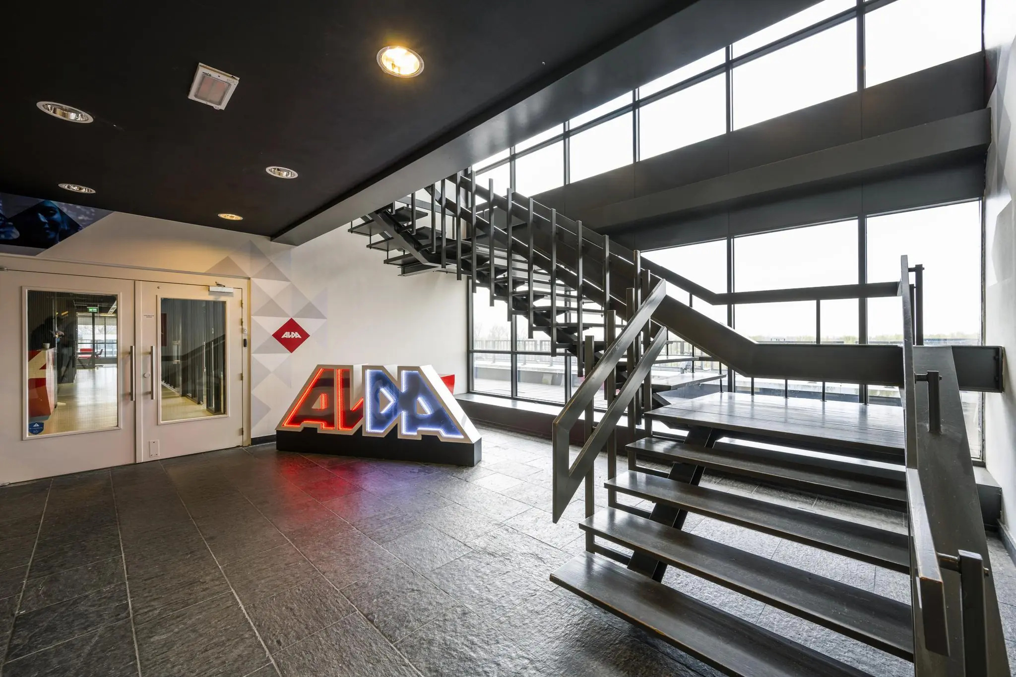 Moderne entreehal met stalen trap, grote raampartijen en een verlicht ALDA-logo bij Anthony Fokkerweg.
