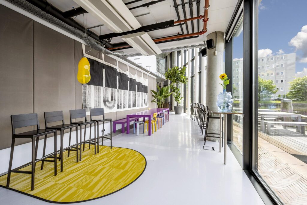 Moderne, kleurrijke ontmoetingsruimte aan de Generaal Vetterstraat met hoge ramen, designmeubilair en uitzicht op een stadsgebouw.