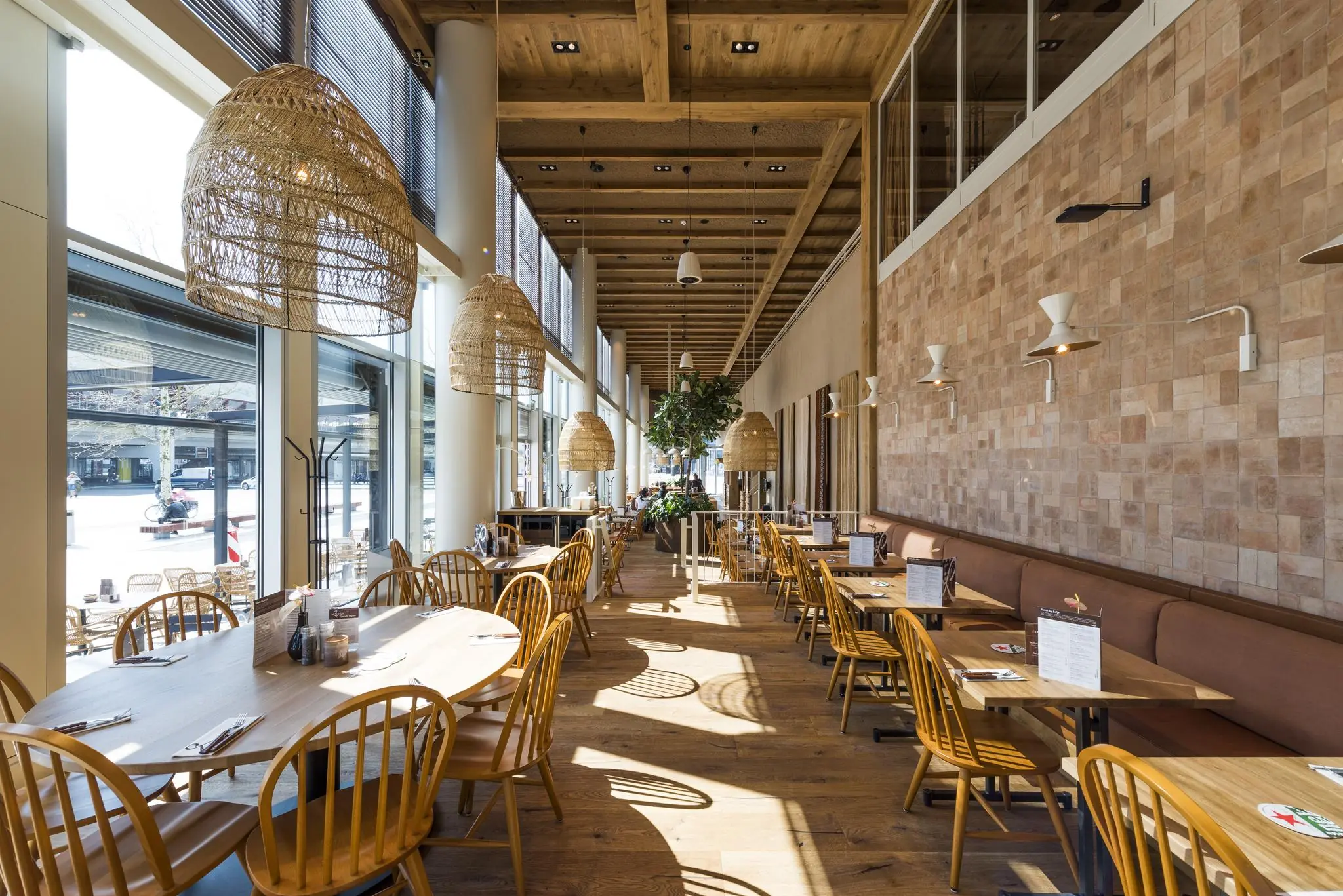 Modern en ruim restaurantinterieur aan het Mandelaplein met houten meubels, grote ramen en hangende rieten lampen.
