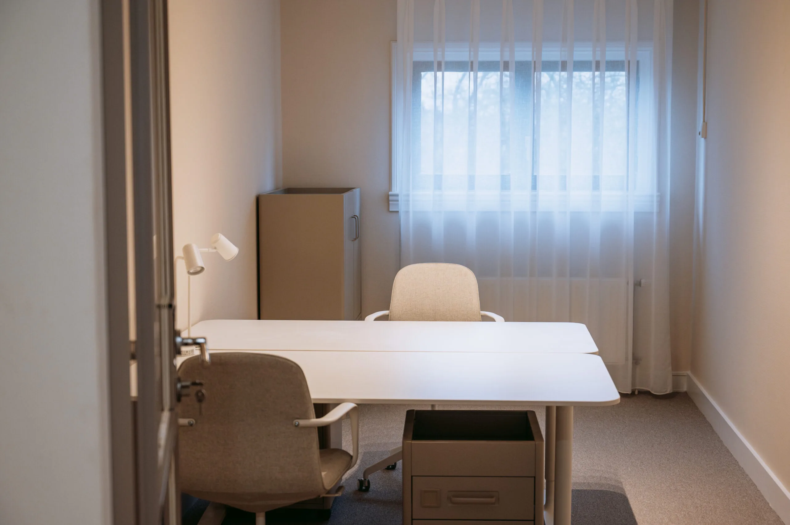 Kleine, minimalistisch ingerichte kantoorruimte met twee stoelen, een bureau, een ladeblok en zacht invallend daglicht door een gordijn.