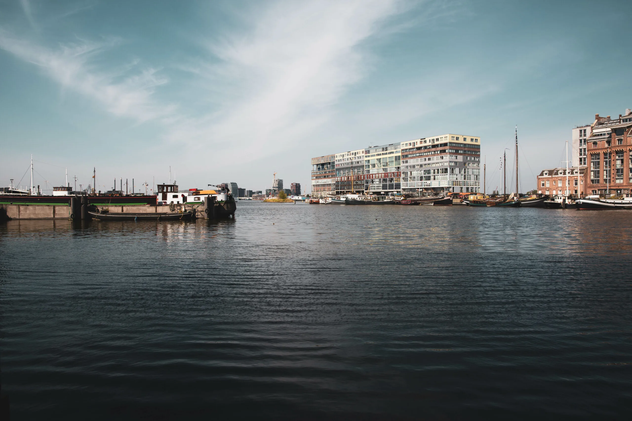 Uitzicht over het water op de Houthavens in Amsterdam met het moderne HNK-gebouw op de achtergrond en aangemeerde boten op de voorgrond.