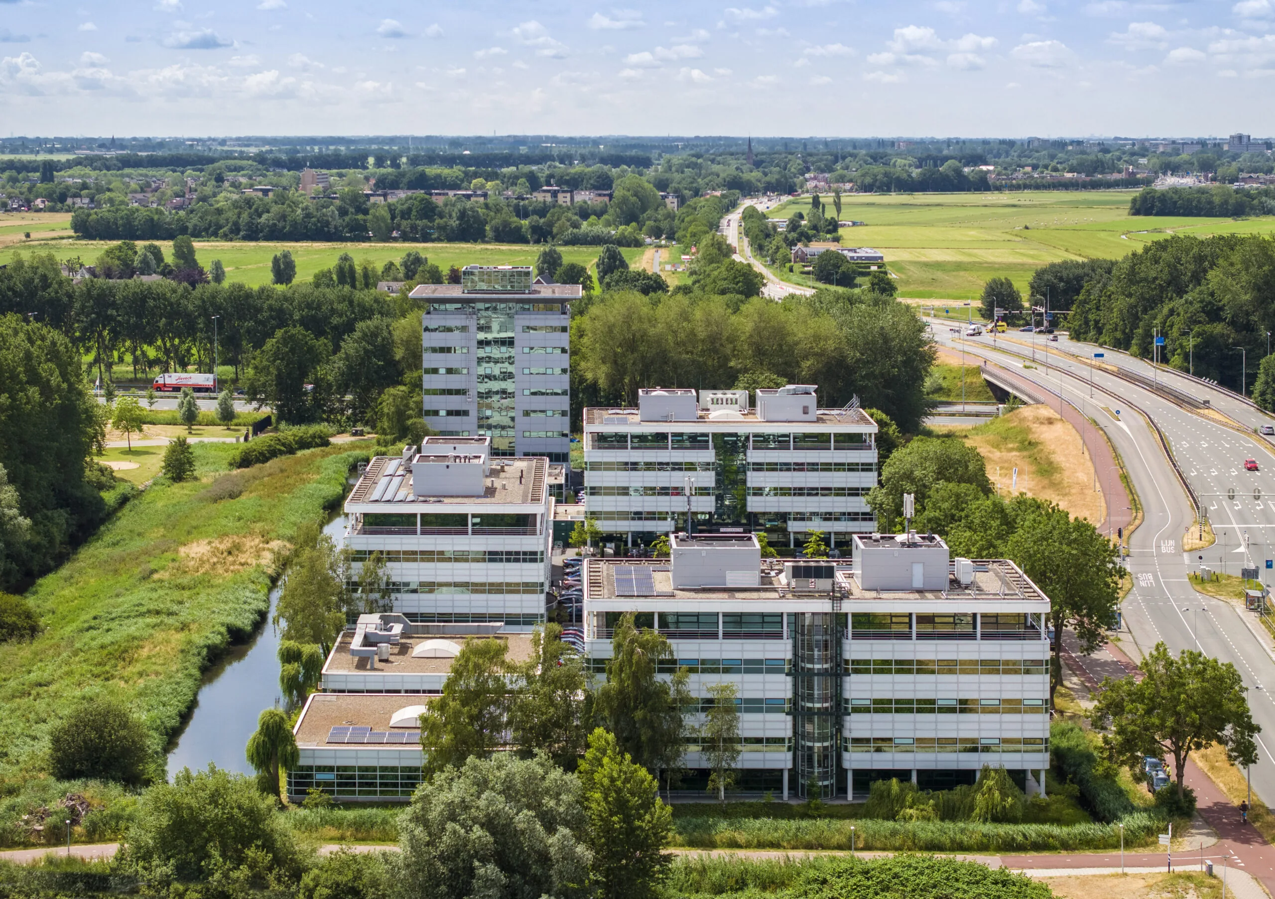 Luchtfoto van een modern kantoorgebouwcomplex omringd door groen, wegen en weilanden.