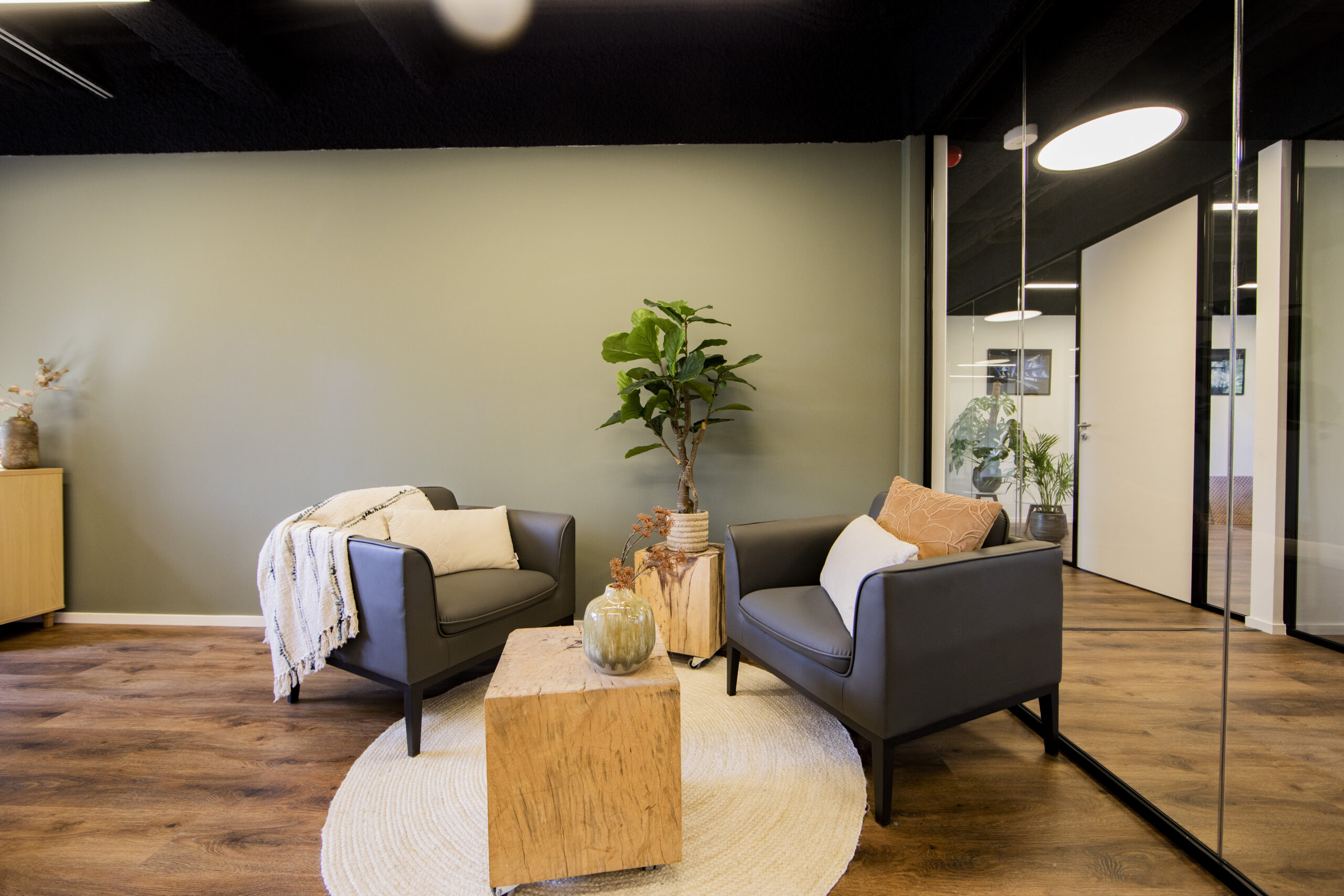 Twee comfortabele fauteuils met kussens naast een houten bijzettafel en planten in een modern kantoor INTERIEUR in Woerden.