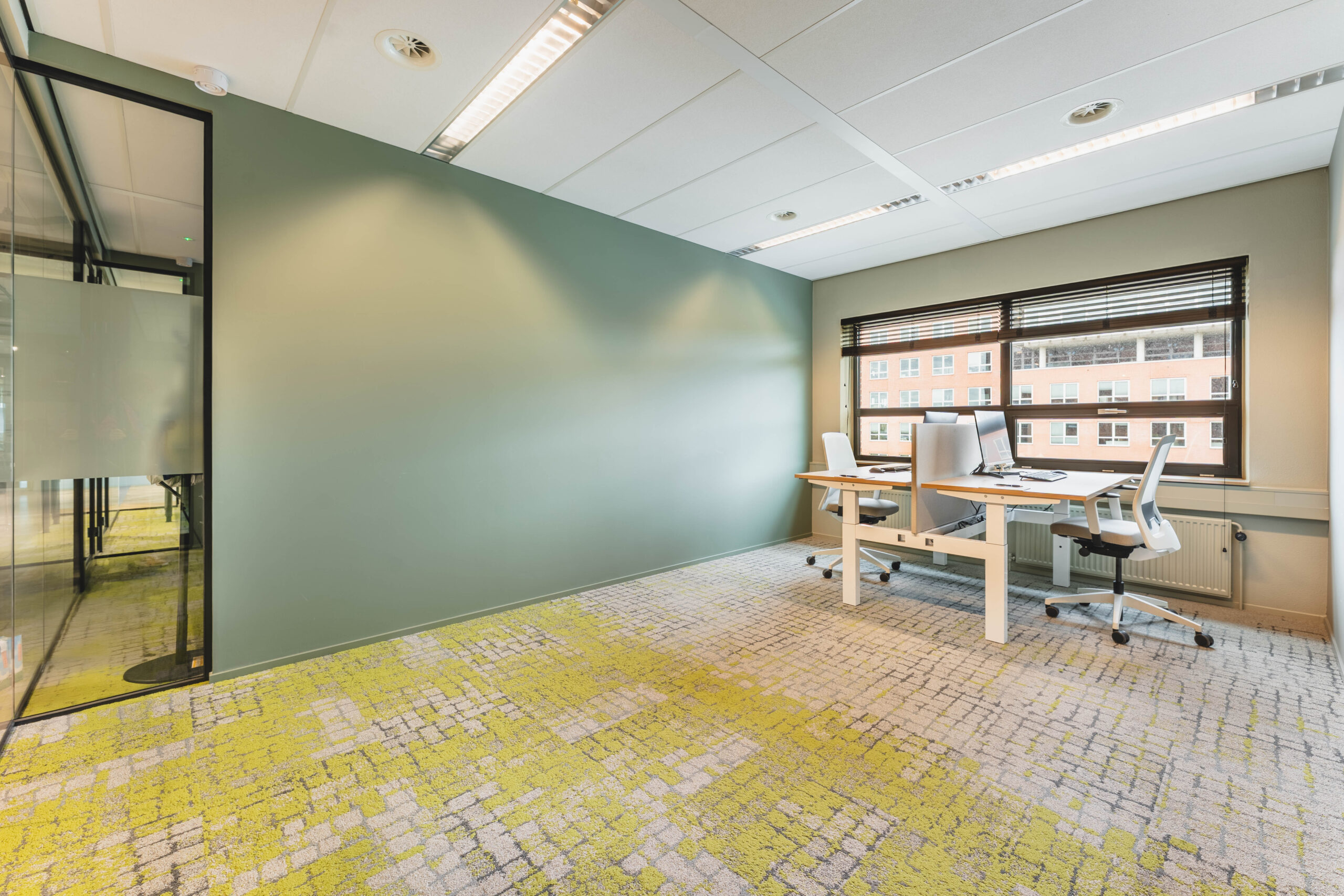 Het toont een moderne kantoorruimte met een groene muur, glaswand en bureau met twee stoelen bij een raam.