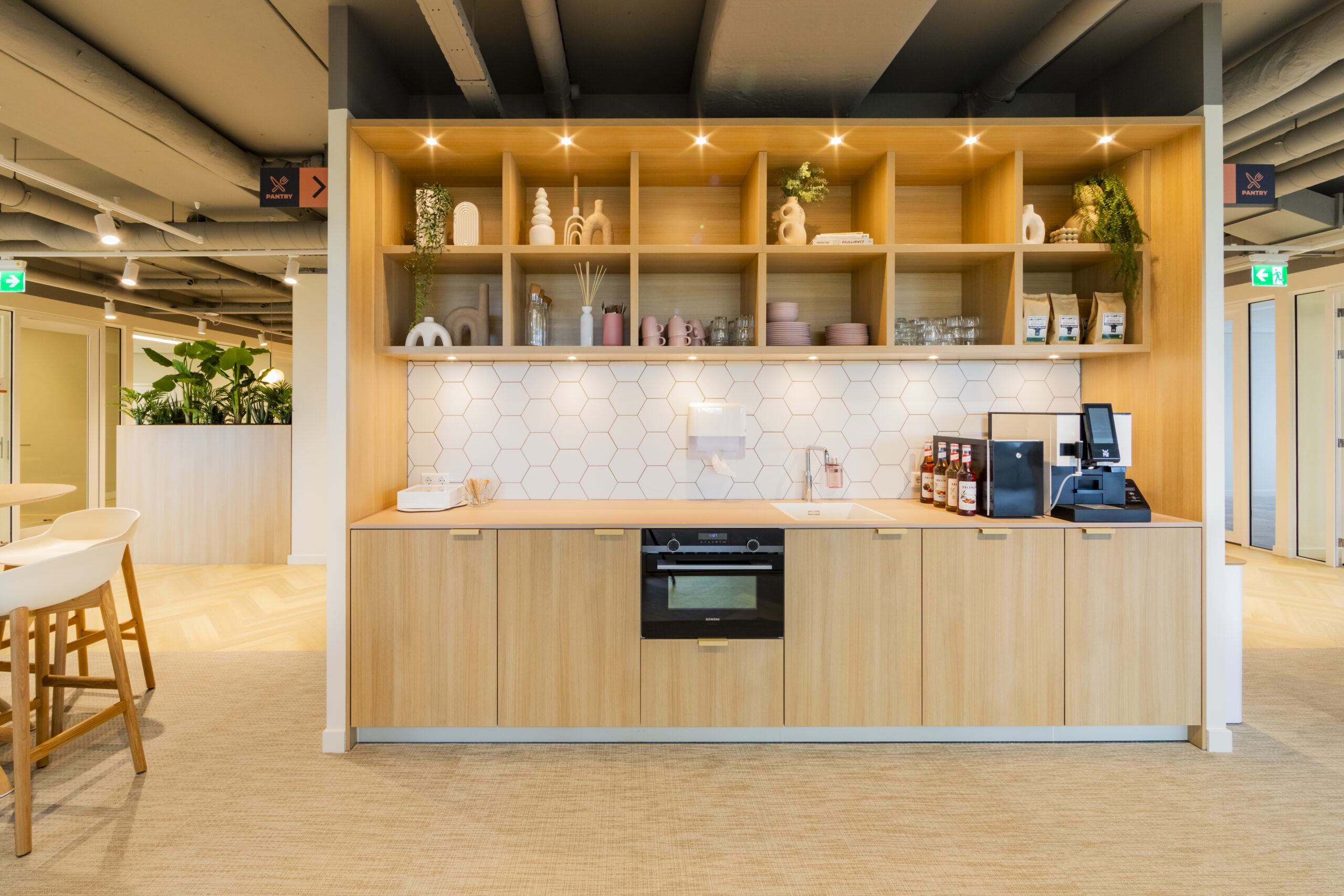 Moderne pantry met houten kasten, planten en keukengerei, onder een plafond met zichtbare leidingen.