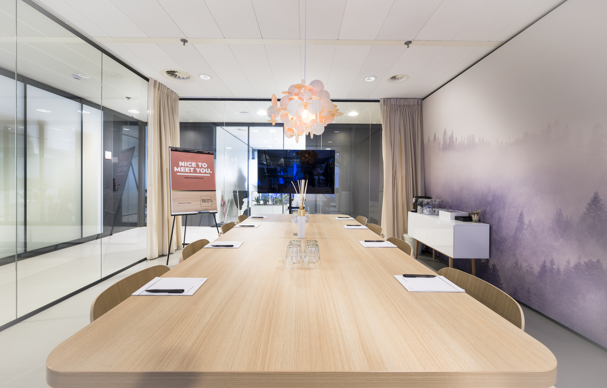 Een moderne vergaderruimte met een lange houten tafel, sfeerverlichting en een muur met boslandschap.