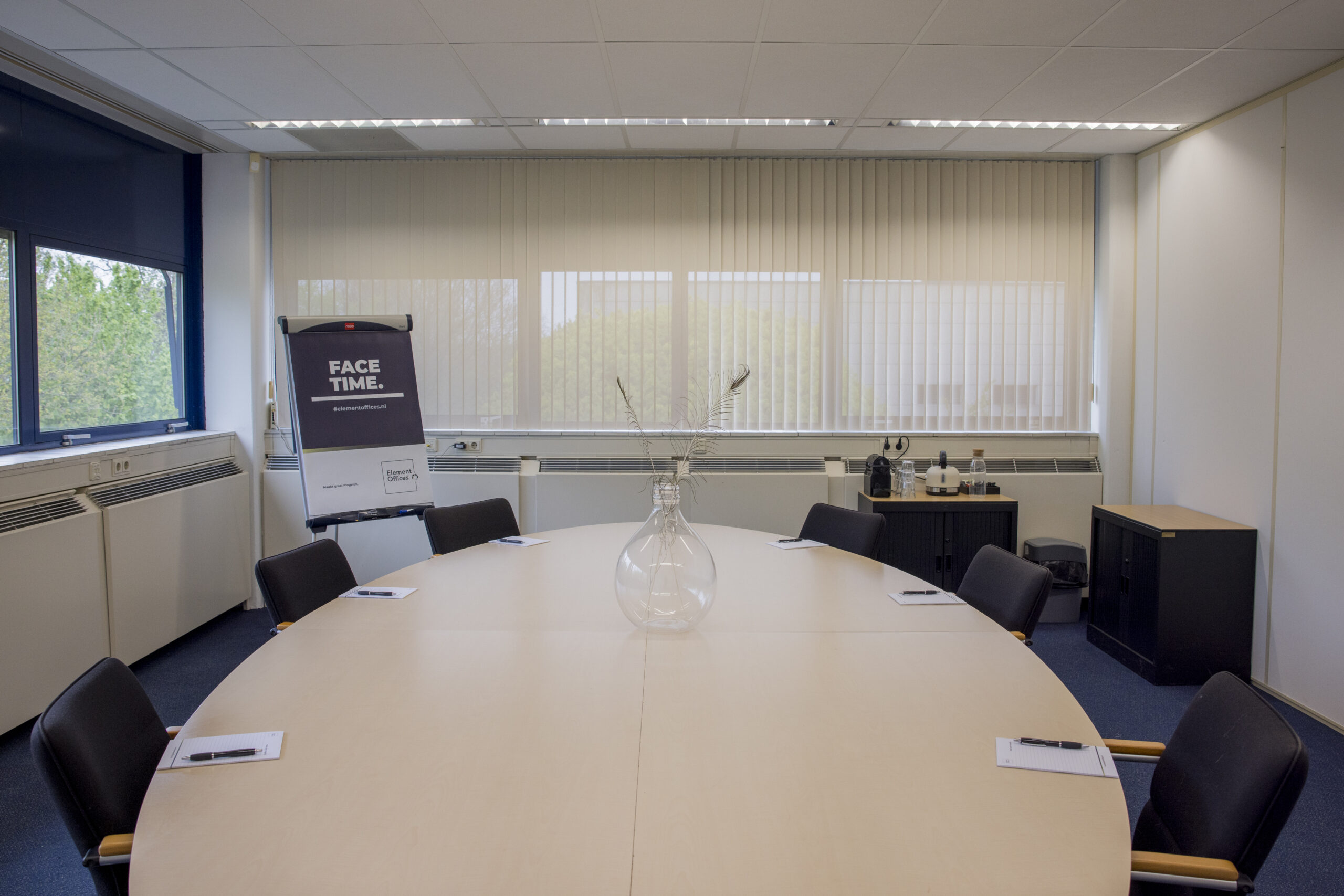 Een vergaderruimte met ovale tafel, stoelen, flipover met de tekst "FACE TIME" en een vaas met veren.