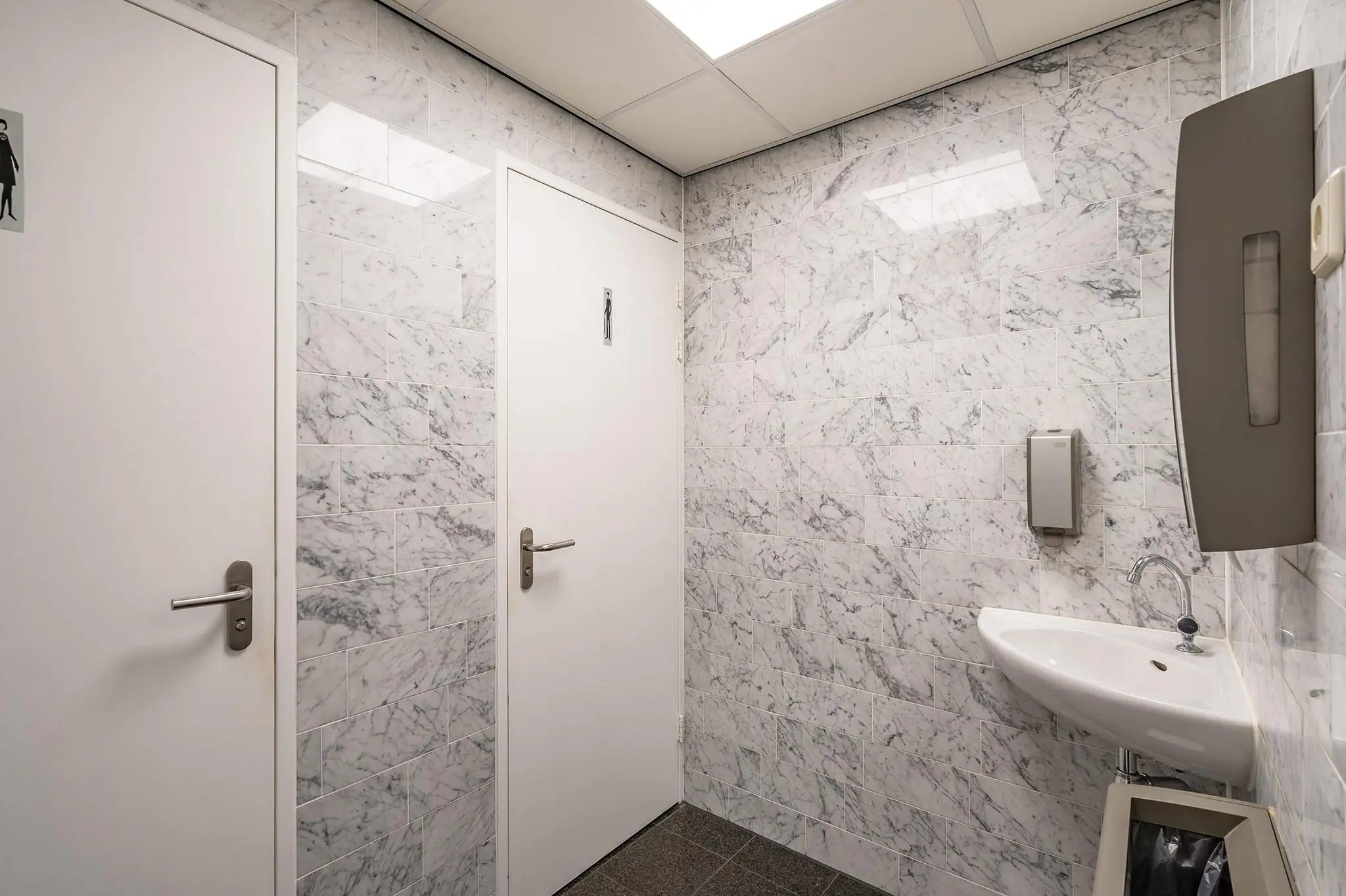 Toiletruimte met marmeren muren, een wastafel, twee deuren en sanitaire voorzieningen in gebouw Rhijnspoor.
