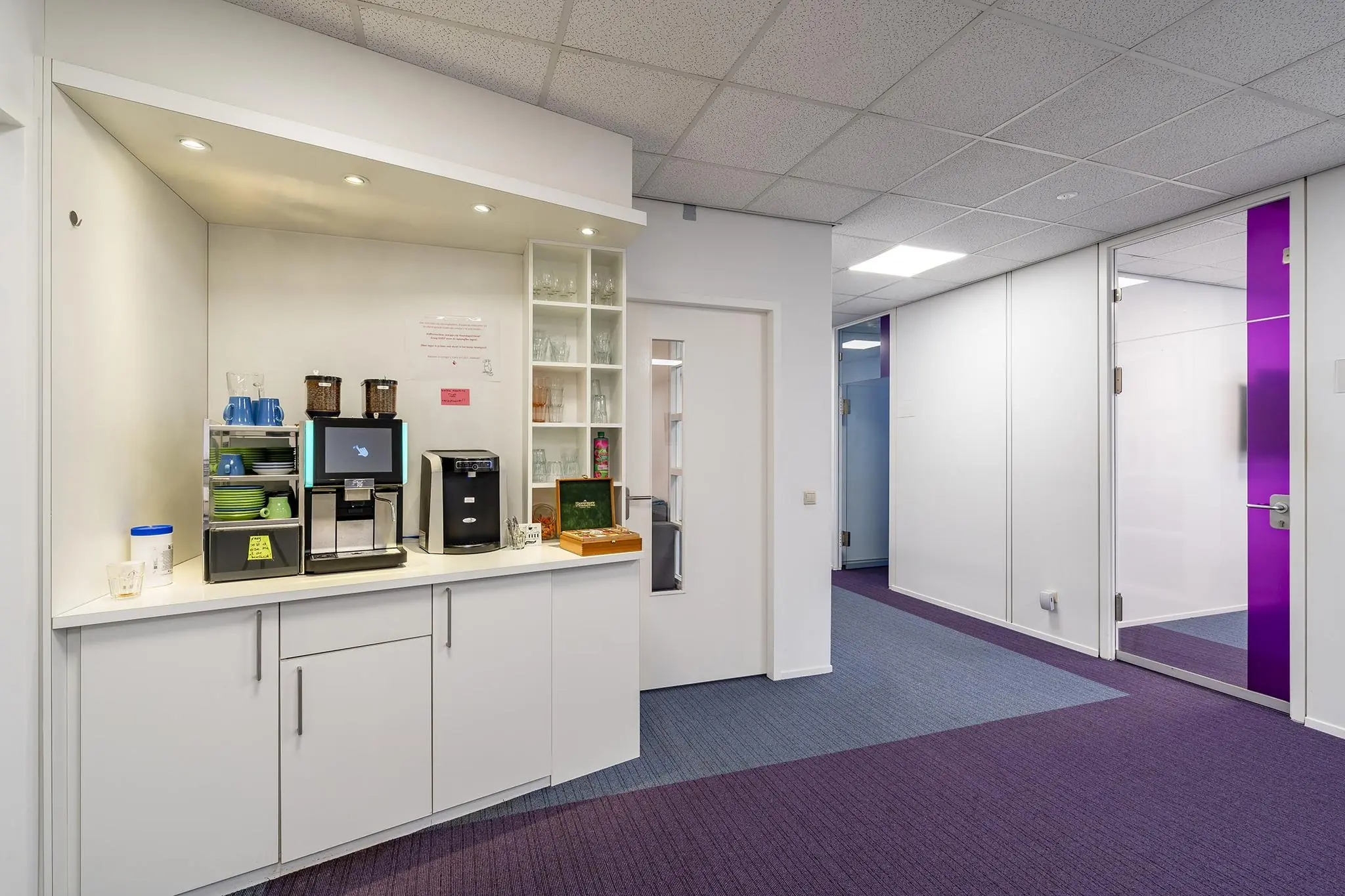 Moderne kantoorpantry met koffiezetapparaat, servies en zicht op een gang met paarse vloerbedekking in het Rhijnspoor-gebouw.
