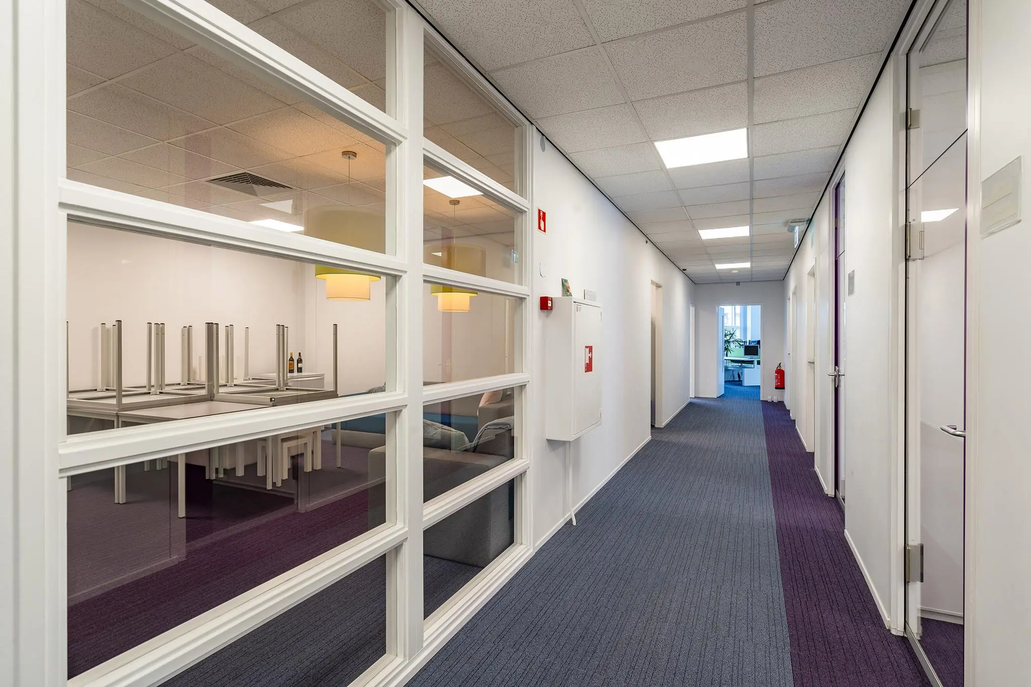 Hal met paarse vloerbedekking en glazen wand met zicht op een kantineruimte met opgestapelde stoelen in Rhijnspoor-gebouw.