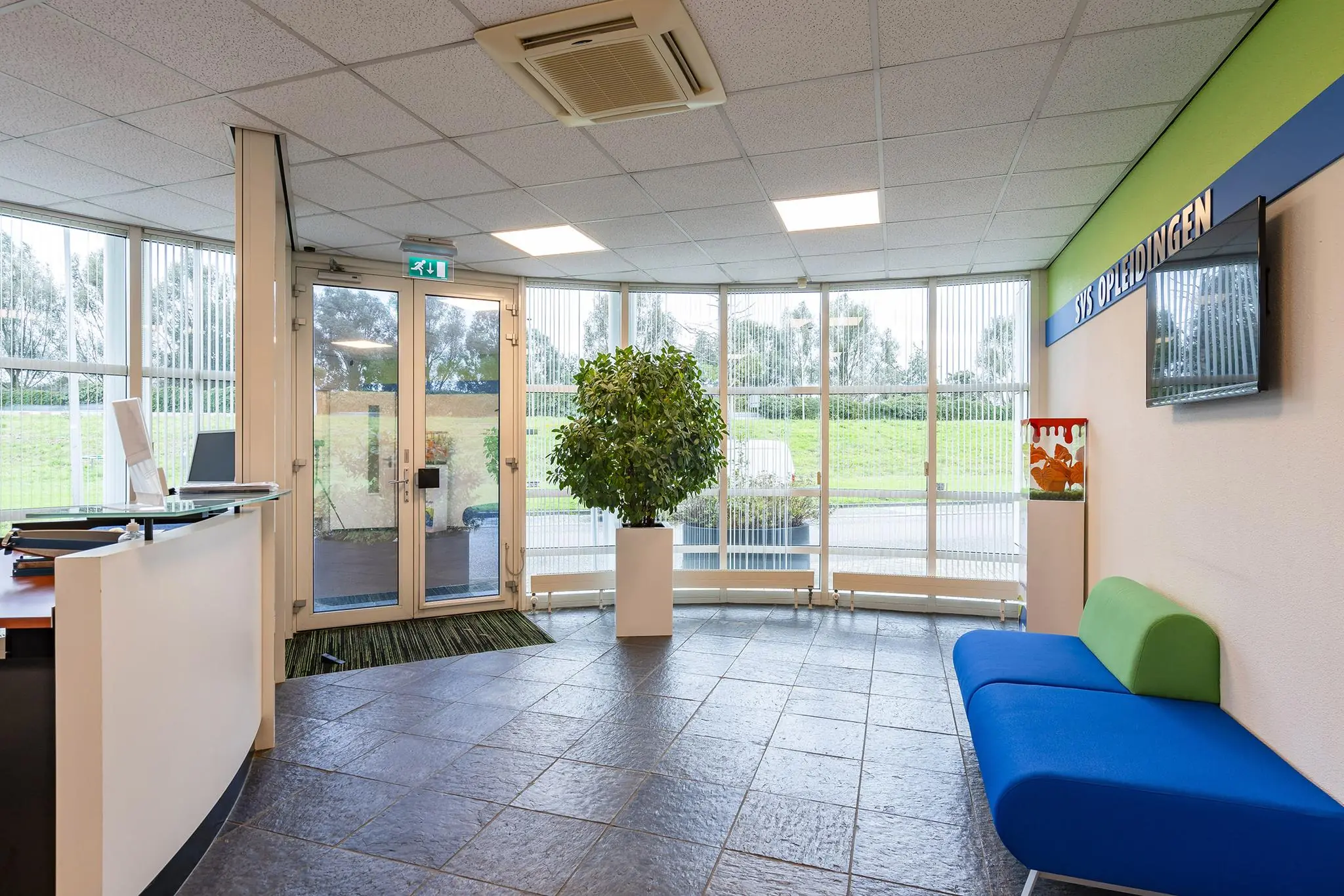 Moderne ontvangstruimte van het gebouw Rhijnspoor met een balie, blauwe bank, grote plant en bord met het logo van SYS Opleidingen.