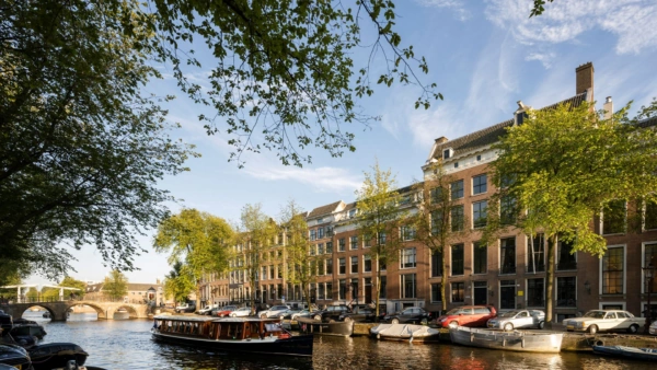 Zicht op de Herengracht in Amsterdam met grachtenpanden, geparkeerde auto's en een rondvaartboot op het water.