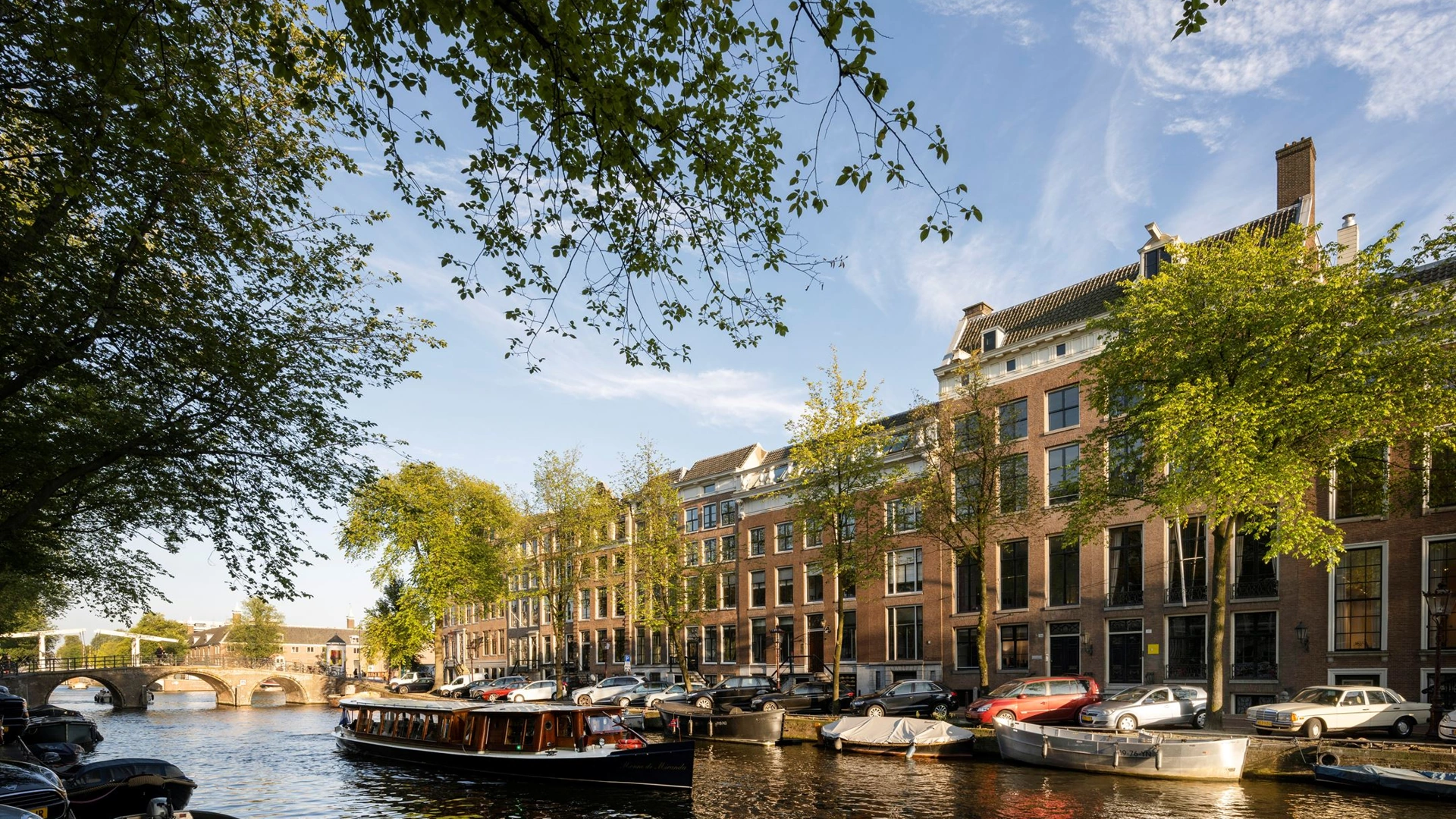 Zicht op de Herengracht in Amsterdam met grachtenpanden, geparkeerde auto's en een rondvaartboot op het water.