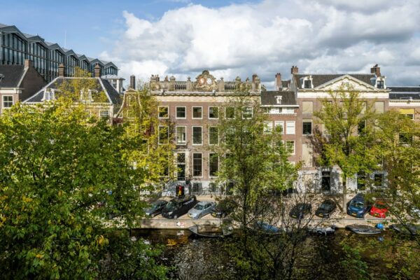 Zicht op monumentale grachtenpanden aan de Herengracht in Amsterdam, gedeeltelijk omgeven door bomen en geparkeerde auto's langs het water.